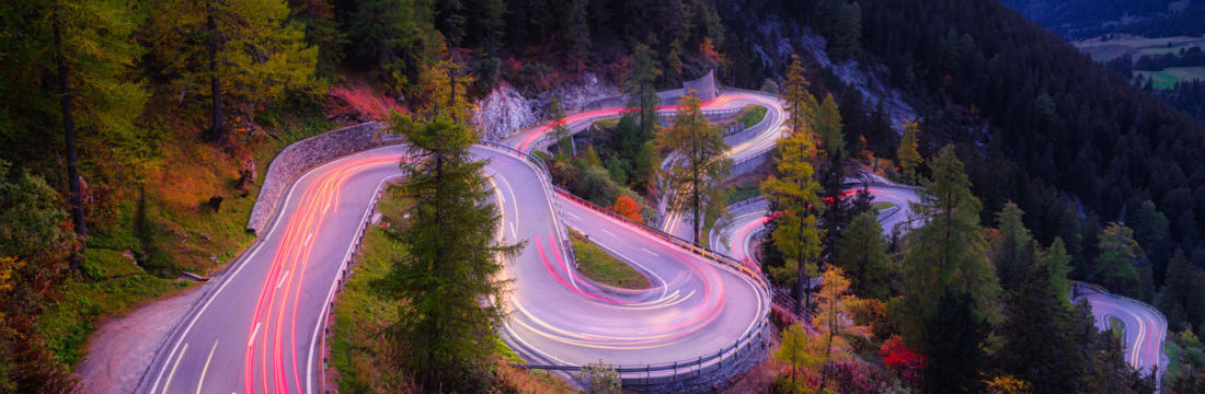 Maloja pass, Switzerland. A road with many curves among the forest. A blur of car lights. Landscape in evening time.